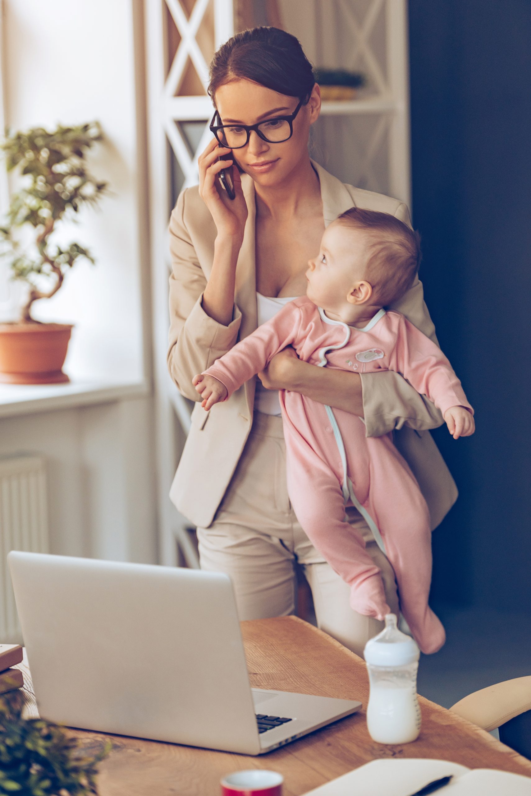 Working mother with baby. Young beautiful businesswoman talking on mobile phone and looking at laptop while standing with her baby girl at her working place